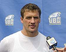 Photo is a profile head shot of Ryan Lochte, a 28-year-old white man with sandy brown hair and blue eyes, standing behind a microphone