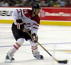 Hockey player in white Canada uniform. He guides a puck across the ice with his stick.