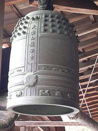 A large, greenish-grey bell hangs from a beamed wooden ceiling