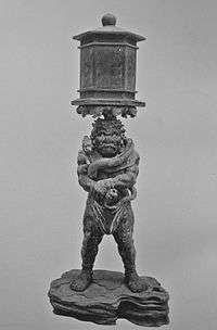 Ryūtōki. Front view of a stocky statue with a demon face. He is balancing a lantern on his head. He is grabbing his right wrist with the left hand. Black and white photograph.