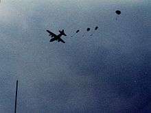 Four small parachutes can be seen coming out the back of a distant, silhouetted plane in flight. It is daytime, however the sky is dark and cloudy.