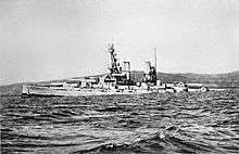 A large light gray battleship tilts back in choppy water as it slowly sinks.