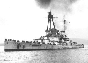 front view of a large warship with a prominent tripod mast above the superstructure at anchor. Two gun turrets are visible and smoke is rising from the two funnels.