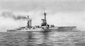 A large gray warship bristling with guns, with a tall mast and two smoke stacks, sits on a calm sea