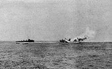 A large, burning warship rolling over and sinking; a smaller, black ship is nearby with two small boats.