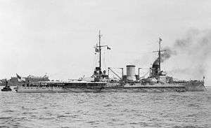 Side view of a gray warship with two masts and two visible gun turrets at anchor. Smoke is rising from one of the two funnels.
