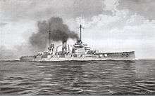 A large gray warship steams through calm seas; thick black smoke pours from two tall smoke stacks