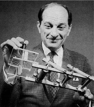 A smiling balding man in a suit and tie holds a strange device that looks like a frame