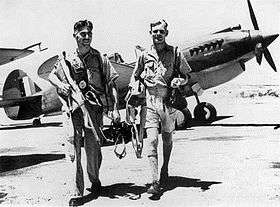 Two men in military uniforms walking among single-engined aircraft on an airfield