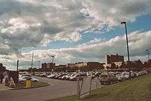 Several rows of cars parked in an outdoor parking lot under a blue sky with billowy clouds.  Many buildings are in the background.