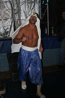 Adult Arab shirtless male wearing a turban, reflective blue pants, and white wrestling boots.
