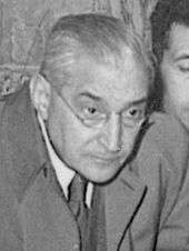 A grey-haired gentleman wearing a suit, an overcoat and glasses leans forward slightly and looks at something out of shot.