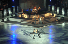 A male and female figure skater spin around each other on the ice while a band plays in the background.