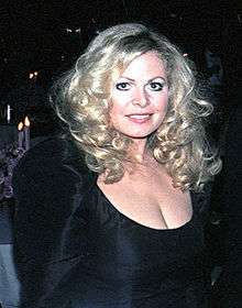 A standing woman with blond hair wearing a black dress looking directly at the camera and smiling.