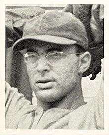 A man wearing glasses and a dark baseball cap with an overlapping "STL" on the front has his hands behind his head as if preparing to throw a ball.