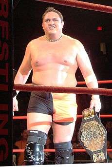 An adult Samoan male wearing orange and black tights with black wrestling boots standing in a wrestling ring with red ropes while holding a black belted championship belt.