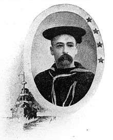 Head and shoulders of a man with a bushy drooping mustache wearing a flat cap and sailor suit. Around the portrait is an oval frame and a depiction of a steamship underway.