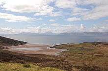 A sandy bay at left and an area of rough grassland at right lie next to a body of water in the middle distance. High hills under white clouds line the horizon.