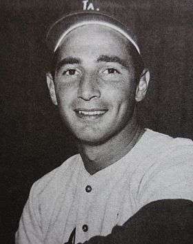 A black-and-white photograph of a smiling man pictured from the chest up; he is wearing a white baseball jersey and dark-colored baseball cap