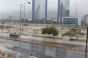 A flooded river in the city of Monterrey runs through the center of the image, and is partially flooding the roadways on both sides of its banks.