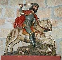 This crudely carved statue shows St James as a bearded knight in armour with a red cloak and upraised sword. He rides a white galloping horse which is trampling the bodies of two armoured men.