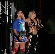 A man and a woman pose together. The dark-haired man is standing on the left and is wearing a blue T-shirt with the Italian flag and the word 'Santino' written upon it, blue wrestling tights, and red kneepads. He is wearing a gold wrestling championship around his waist. The blond haired woman is standing on the right, wearing a black catsuit with the sides of the torso removed to bare part of her midriff. She is holding a wrestling championship upon her left shoulder and has her right arm outstretched.