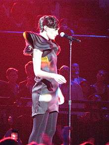 33-year-old Blasko is shown in a three-quarter shot and right profile. She is looking over the top of the microphone on its stand. Her dress has a dark background with coloured parts interspersed. Her right arm is crooked at the elbow. A small tattoo is partly visible on her right upper arm.