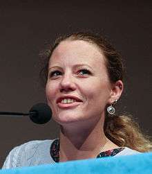 Sarah Harrison, with earrings and long, dirty blonde ponytail, faces left towards a microphone