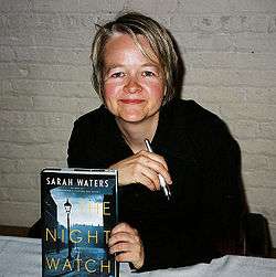 Image of a woman with short blond hair seated at a table holding a pen and a book cover to the camera