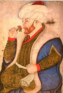 A corpolent bearded young man holding a rose and wearing a turban