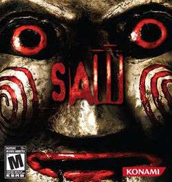 The box art features the face of an antique-looking Billy doll. The face is gold in color and is shiny. It has red eyes and red lips. The word "Saw" in red letters is in the center.