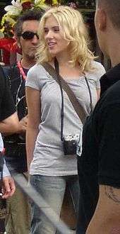 A young woman with tousled medium length blonde hair loosely around her shoulders and face, looking to her right, her right arm slightly behind her, stands in the middle of a group of men. She is dressed in denim jeans, a light gray short sleeved shirt and a camera hangs around her neck.