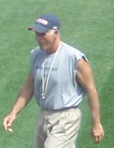 Candid photograph of Scarnecchia walking on a football field wearing a grey sleeveless t-shirt and a blue baseball cap