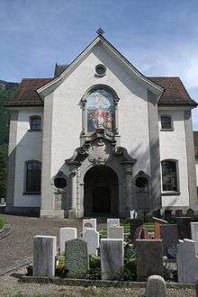 A baroque-style church, white walls with tile roof, and a set of grave stones in front of it.