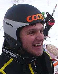 A smiling young man is geared up with a yellow-striped black winter jacket, black and gray protective helmet with orange lettering at the front, and winter goggles around his neck. He is at a snow-covered site along with other people.