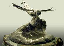 A trophy with a bronze coloured base, above the base is a wave of silver coloured metal. Atop the wave is a silver winged figurine kissing another figurine partly submerged in the wave.
