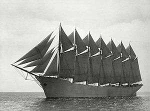 A seven-masted schooner outfit with sails sits high in the water