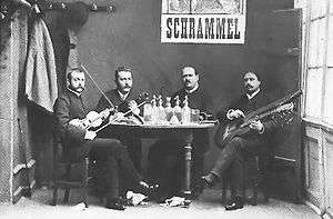 Four musicians with instruments are seated at a table as they face the camera. The lower section of a poster appears on the back wall, displaying the name "Schrammel."