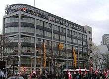A seven-story, modern building, predominantly grey and white, with a cross-like symbol and large letters spelling "Scientology Kirche" at the top. There is a crowd of people in front of the building, some of them with flags; the building itself is decorated with two yellow lengths of cloth and a large yellow cloth flower. Several dozen people are visible on the roof terrace.