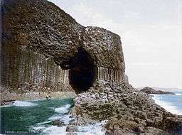 A large dark cave mouth sits in a cliff face, the lower half of which is columnar in appearance. Waves lap the cave entrance.