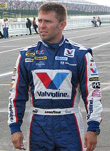 A man in his thirties with a head full of hair. He is wearing blue and white racing overalls with sponsor's logos.