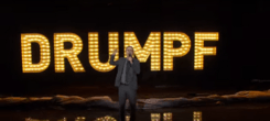 Comedian John Oliver atop a stage, wearing a suit and holding a microphone; in the background is the text "Drumpf"