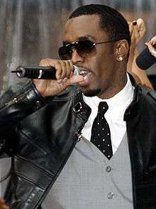 An African-American man, dressed in sunglasses, a white shirt, a black tie and black jacket, performs into a microphone.