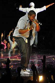 Sean Kingston wearing jeans and a grey shirt, singing on stage.