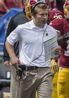 Photograph of McVay on a football sideline wearing a white Washington Redskins polo shirt, khaki pants and a headset and holding a football play sheet in his left hand