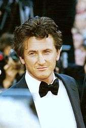 An American man with flowing dark hair, looking towards the camera, wearing a suit