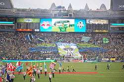 A stand of a stadium with fans holding a large banner depicting the Space Needle. Green and blue flags wave throughout the stand and the flags of different nations are held by people on the field.