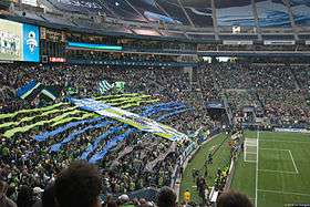 Fans waving flags and unfurling a large green and blue banner behind a goal.