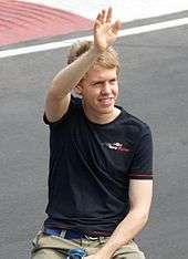 Head, arms and torso of a man in his early twenties with slightly tousled blond hair. He is waving with his right hand. He is wearing a black T-shirt emblazoned with the Scuderia Toro Rosso on the left breast and beige trousers or shorts held up by a blue belt.