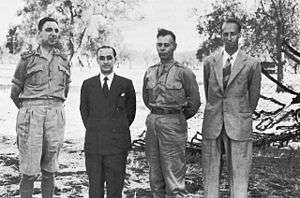 Four men pose awkwardly for a photograph. Two are in shirt sleeve uniforms and the other two are wearing suits. All are bare-headed.